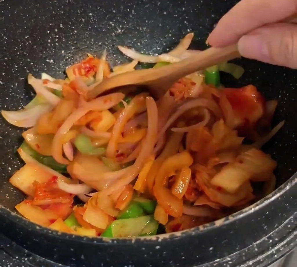 Una cuchara de madera mezclando kimchi de color naranja vibrante con la base de cebollas salteadas.