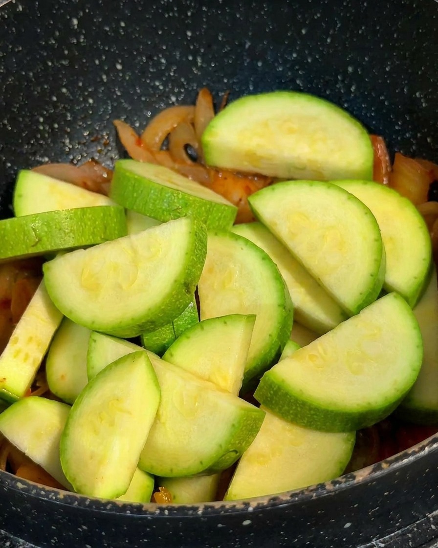 Rodajas de calabacín verde fresco apiladas en una olla sobre una base de kimchi y cebollas cocidas.