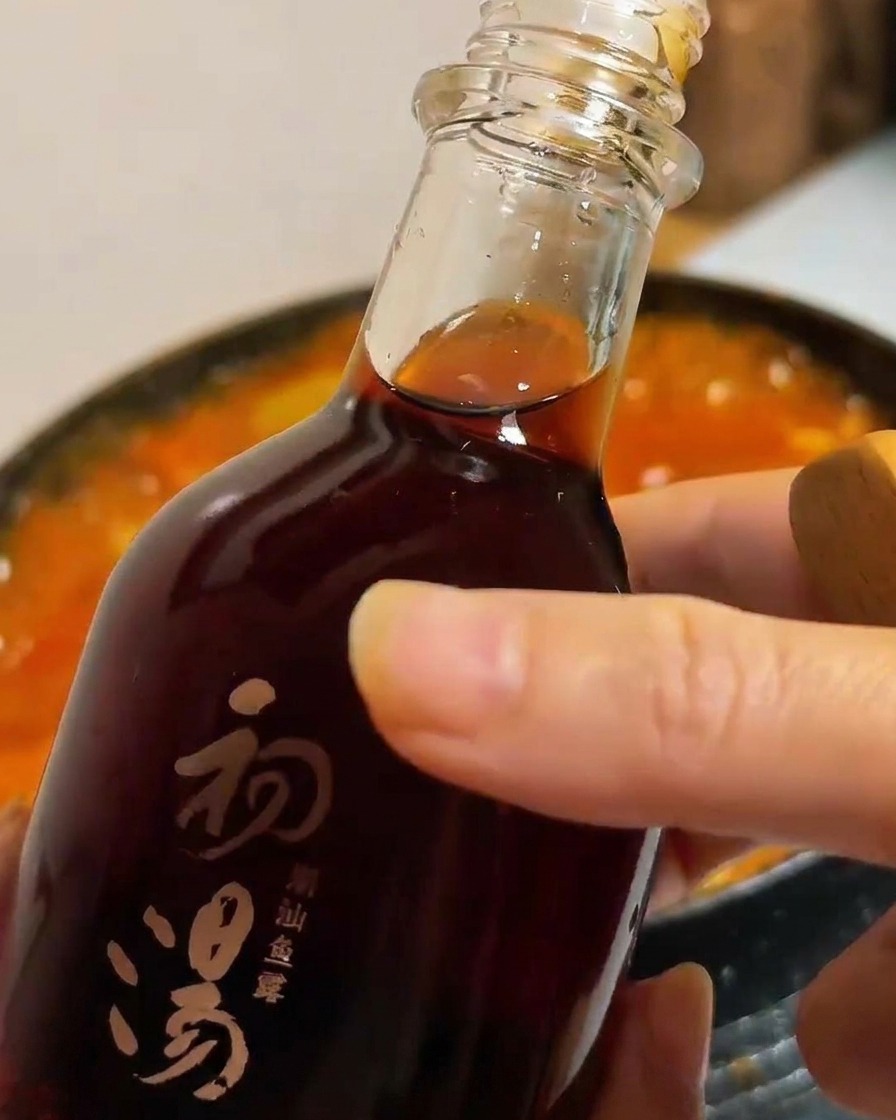Una botella de vidrio oscura vertiendo salsa de pescado en una olla de estofado de tofu y vegetales.