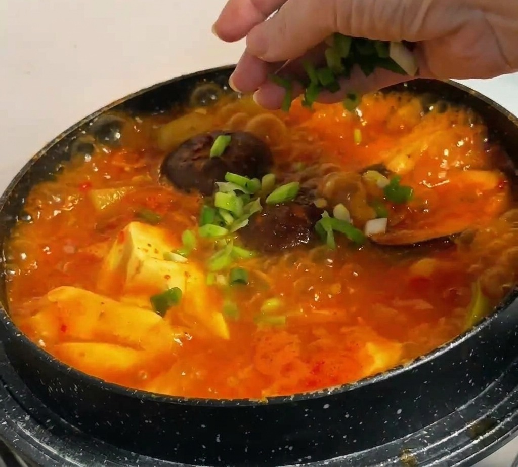 Cebolletas verdes picadas siendo espolvoreadas a mano sobre un estofado coreano rojo vibrante con tofu y setas.
