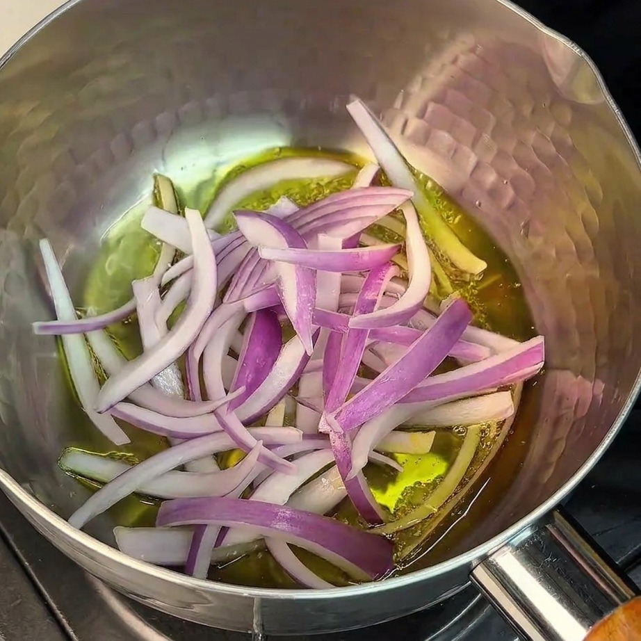 Cebolla morada en rodajas finas sofriéndose en aceite al fondo de una olla de acero inoxidable.