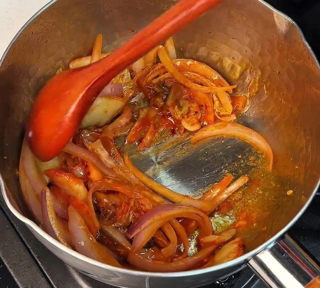 Una cuchara de madera mezclando kimchi picado y cebolla morada en una olla metálica caliente.