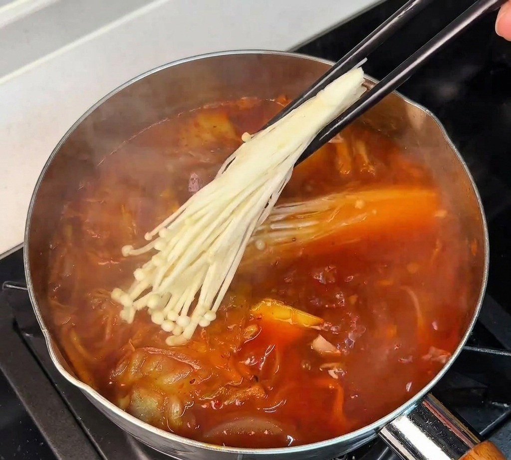 Una mano usando palillos para sumergir un manojo de hongos enoki blancos en una olla de sopa de kimchi hirviendo.