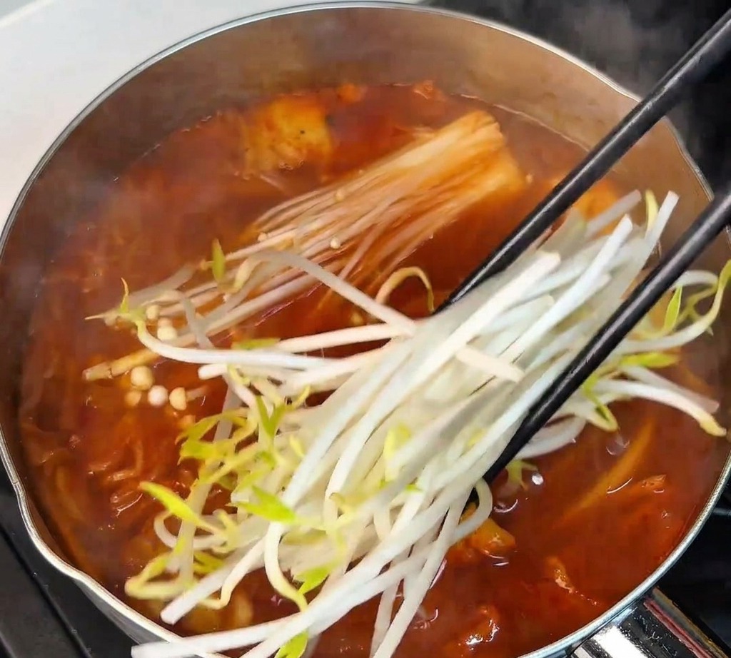 Brotes de soja blancos frescos siendo añadidos con palillos en la olla de sopa roja junto a los hongos enoki.