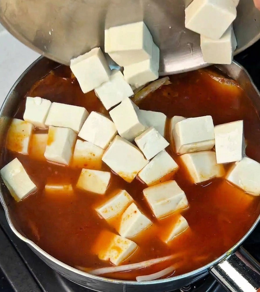 Varios cubos de tofu blanco vertiéndose desde un bol de acero en una olla de caldo de kimchi rojo burbujeante.