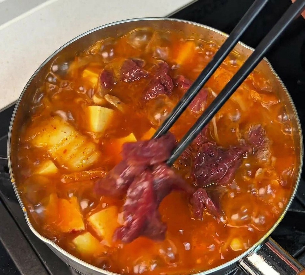 Láminas de ternera cruda marinada siendo colocadas en una sopa roja burbujeante con palillos negros.