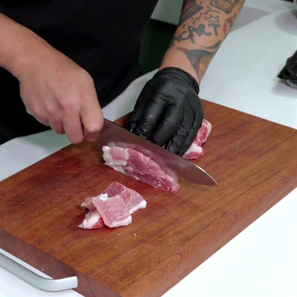 Mano con guante negro usando un cuchillo grande para cortar panceta de cerdo cruda en tiras pequeñas.