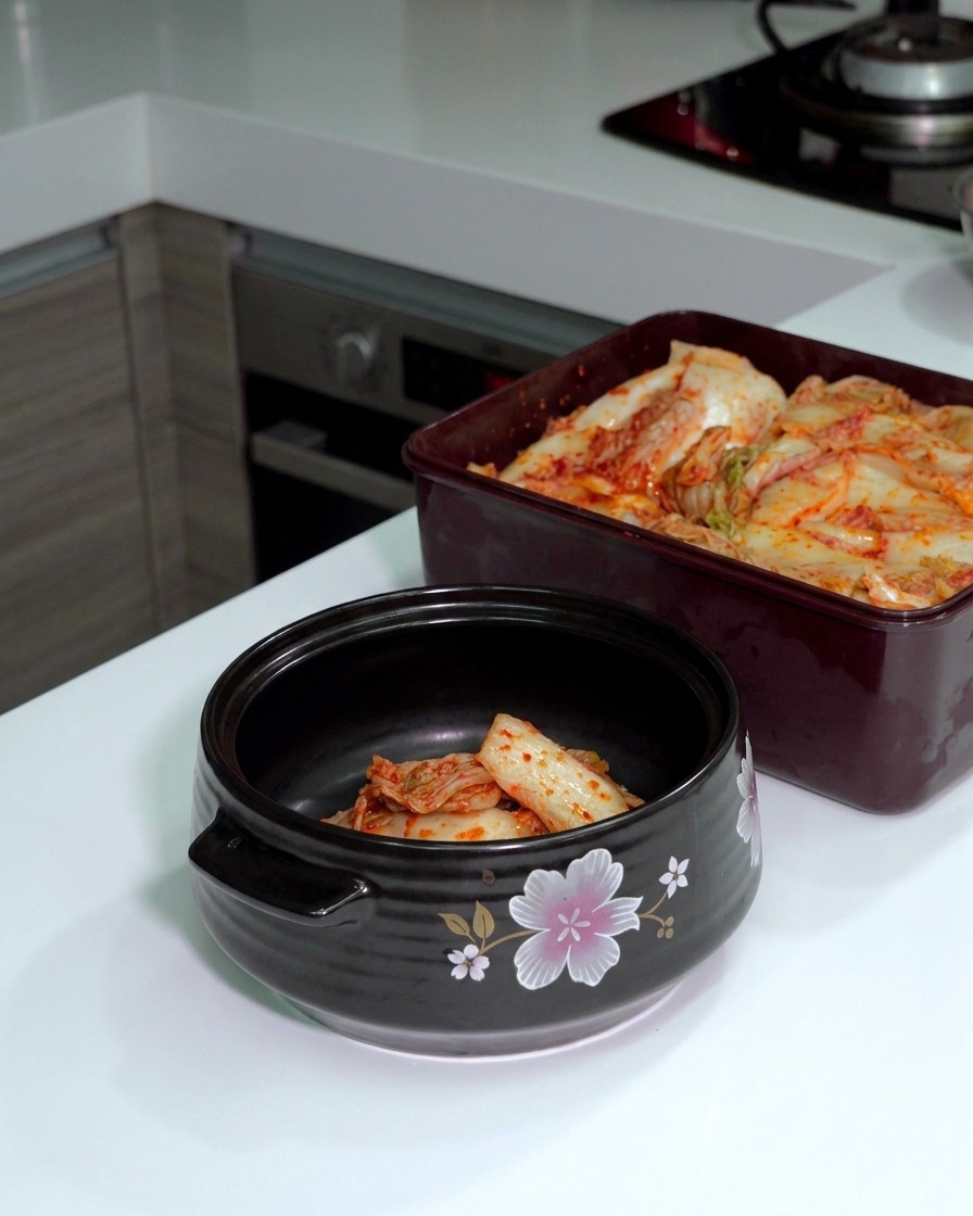 Kimchi de col fermentada siendo cortado con tijeras plateadas dentro de una olla de cerámica oscura.