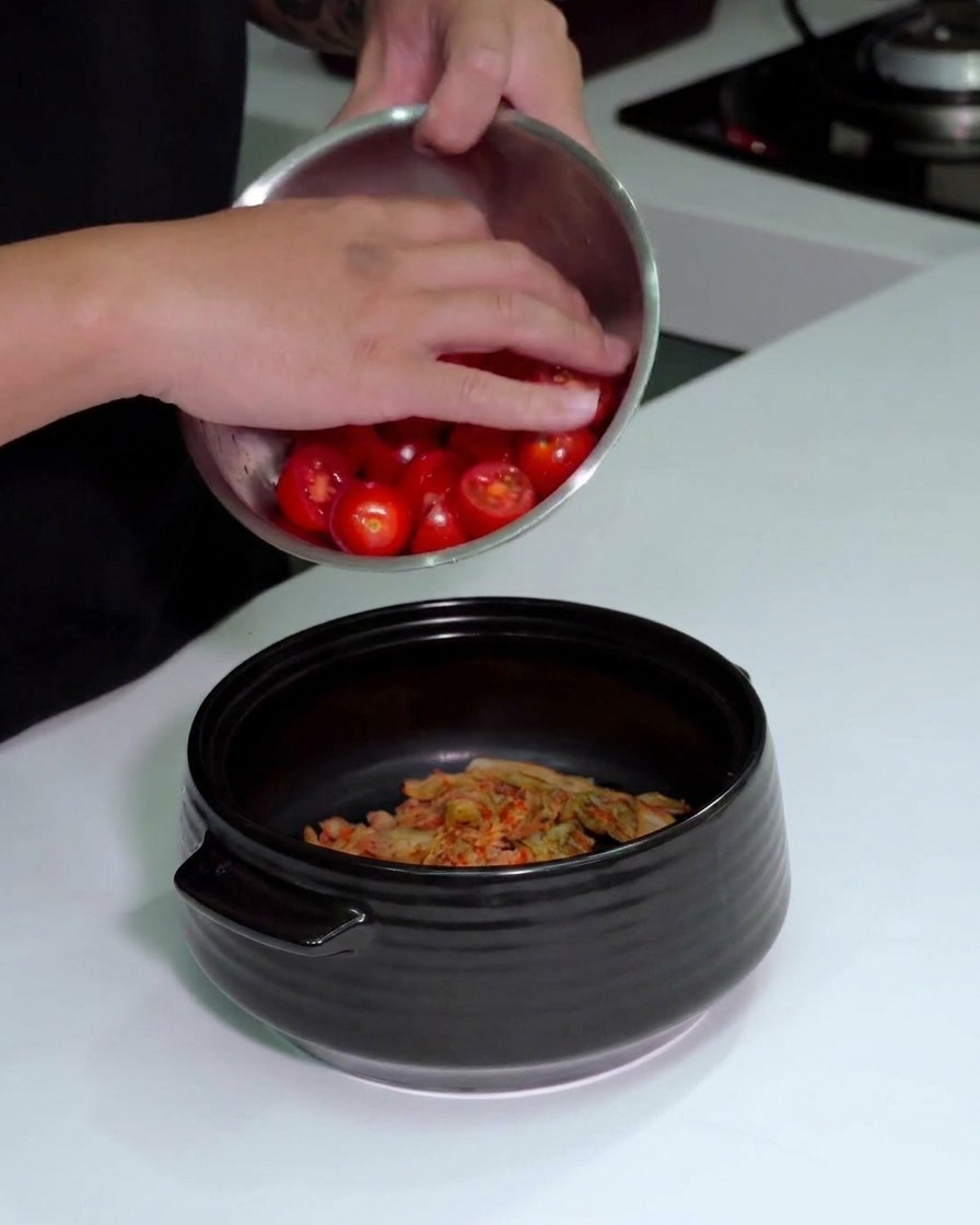 Persona vertiendo un cuenco de tomates cherry cortados en una olla que ya contiene el kimchi picado.