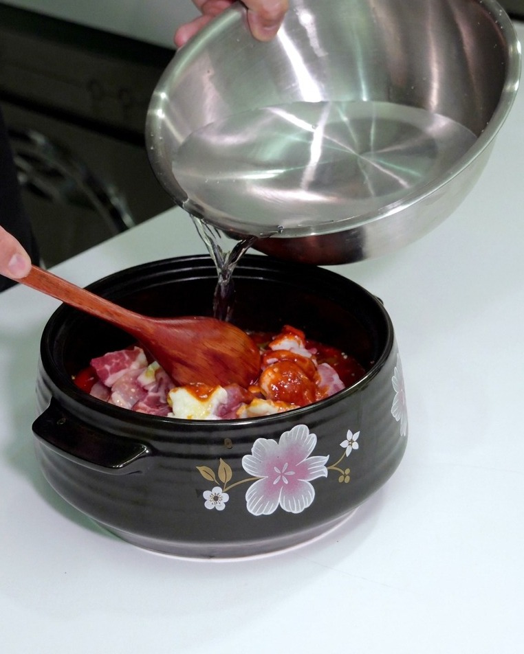 Persona vertiendo agua desde un cuenco a la olla que contiene la carne y verduras sazonadas.