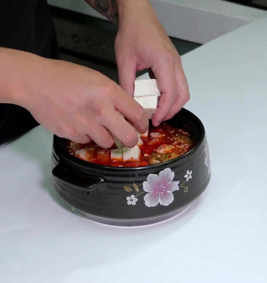 Manos colocando cubos de tofu fresco en la olla llena con la base de sopa de kimchi y panceta.