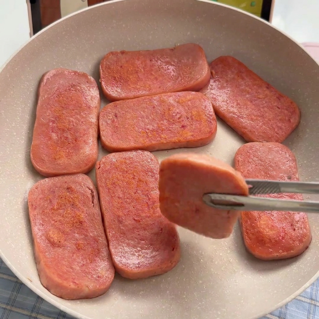 Rodajas rectangulares de carne de almuerzo rosada sellándose en una sartén antiadherente hasta que están doradas.