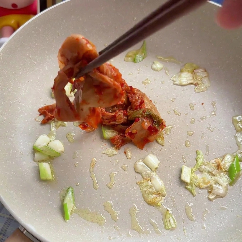 Unos palillos añadiendo una porción de kimchi de col roja troceado a una sartén con vegetales sofritos.