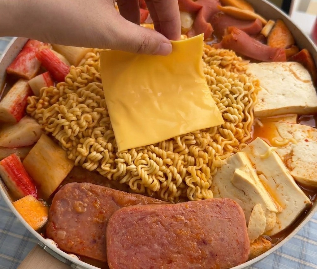 Una mano colocando una loncha cuadrada de queso amarillo sobre los fideos ramen en una olla humeante.