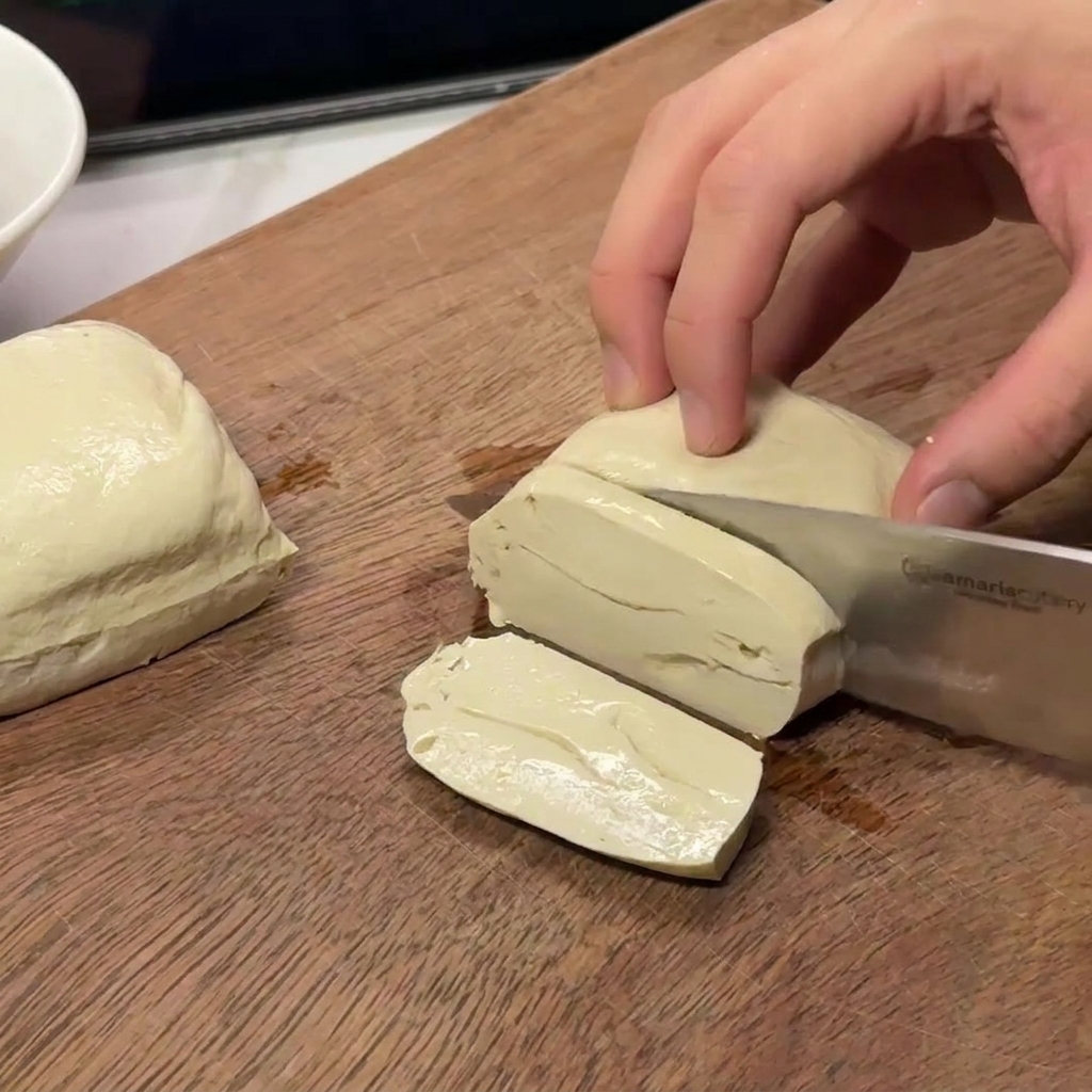 Manos cortando un bloque de tofu blanco firme en piezas rectangulares uniformes.