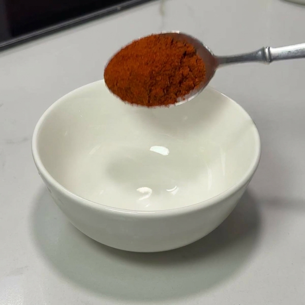 Una cuchara vertiendo chile rojo en polvo en un bol de cerámica blanca.