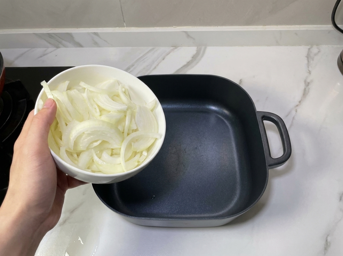 Una mano sosteniendo un bol con cebollas laminadas sobre una olla negra vacía.