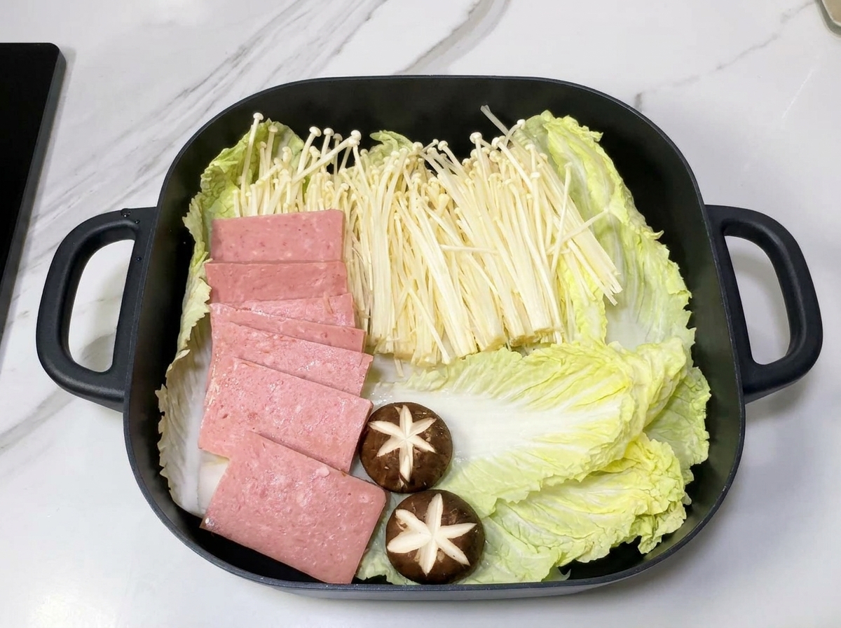 Una olla cuadrada con capas de col china, setas enoki y rodajas de spam dispuestas sobre la cebolla.