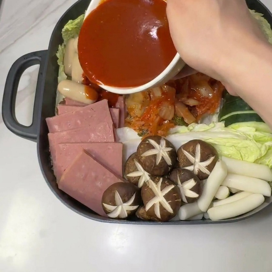 Salsa roja picante de gochujang vertiéndose desde un bol blanco sobre el centro de los ingredientes crudos del estofado.