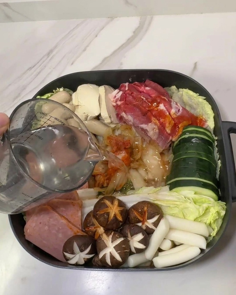 Una jarra medidora vertiendo agua en una olla llena de ingredientes de estofado coreano.