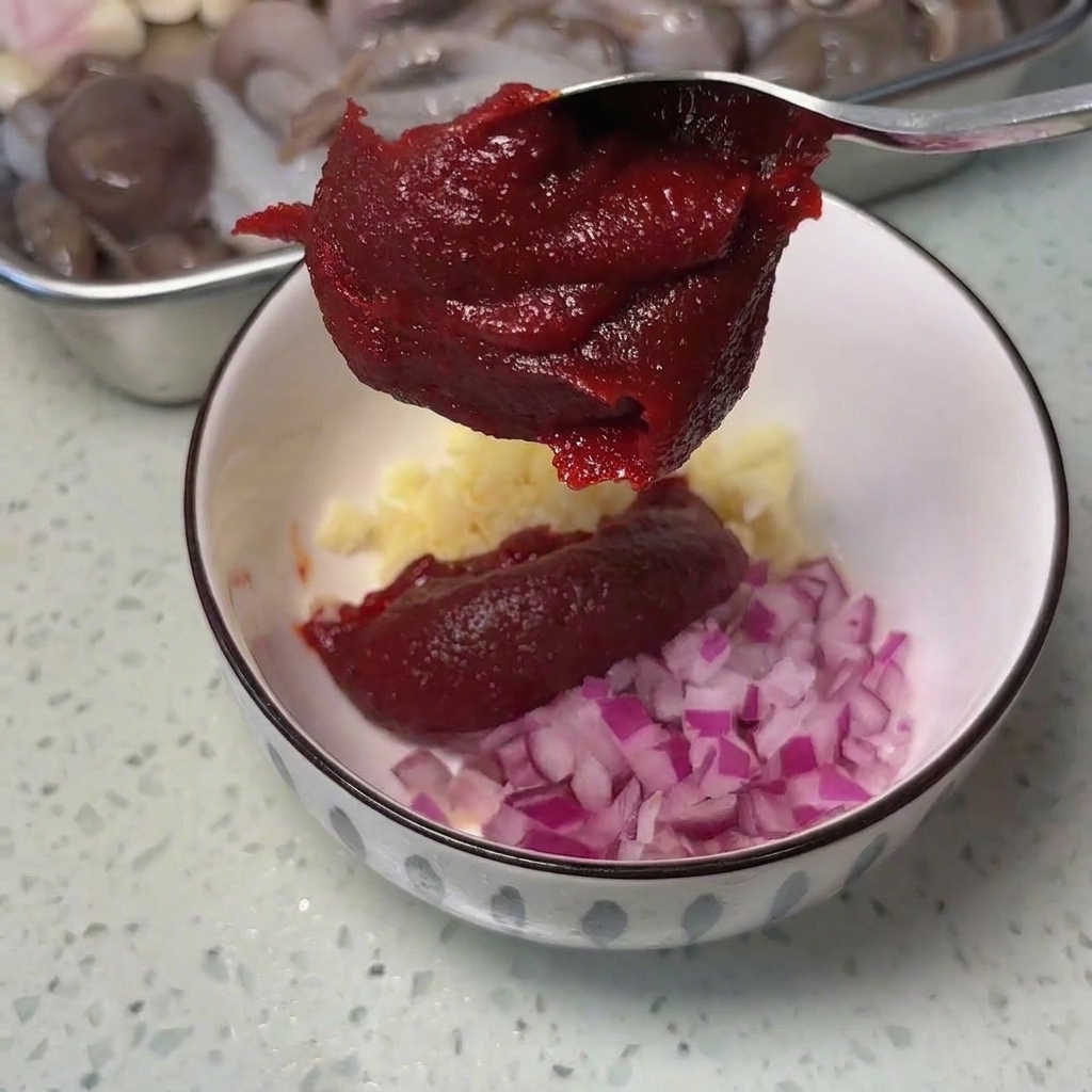 Una cuchara de metal con una porción generosa de pasta roja de chile coreano sobre un cuenco con ajo y cebolla picada.