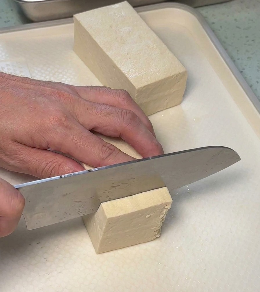 Un cuchillo de cocina cortando un bloque de tofu blanco firme en rectángulos iguales sobre una tabla de cortar.