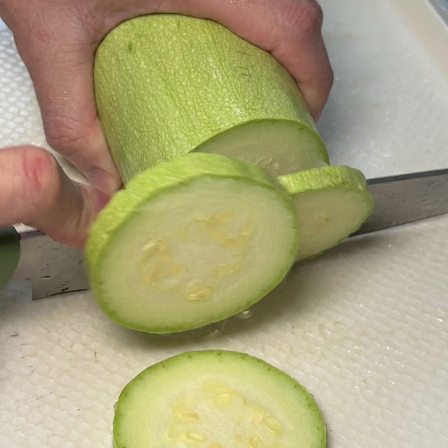 Unas manos cortando un calabacín verde en rodajas circulares sobre una tabla blanca.