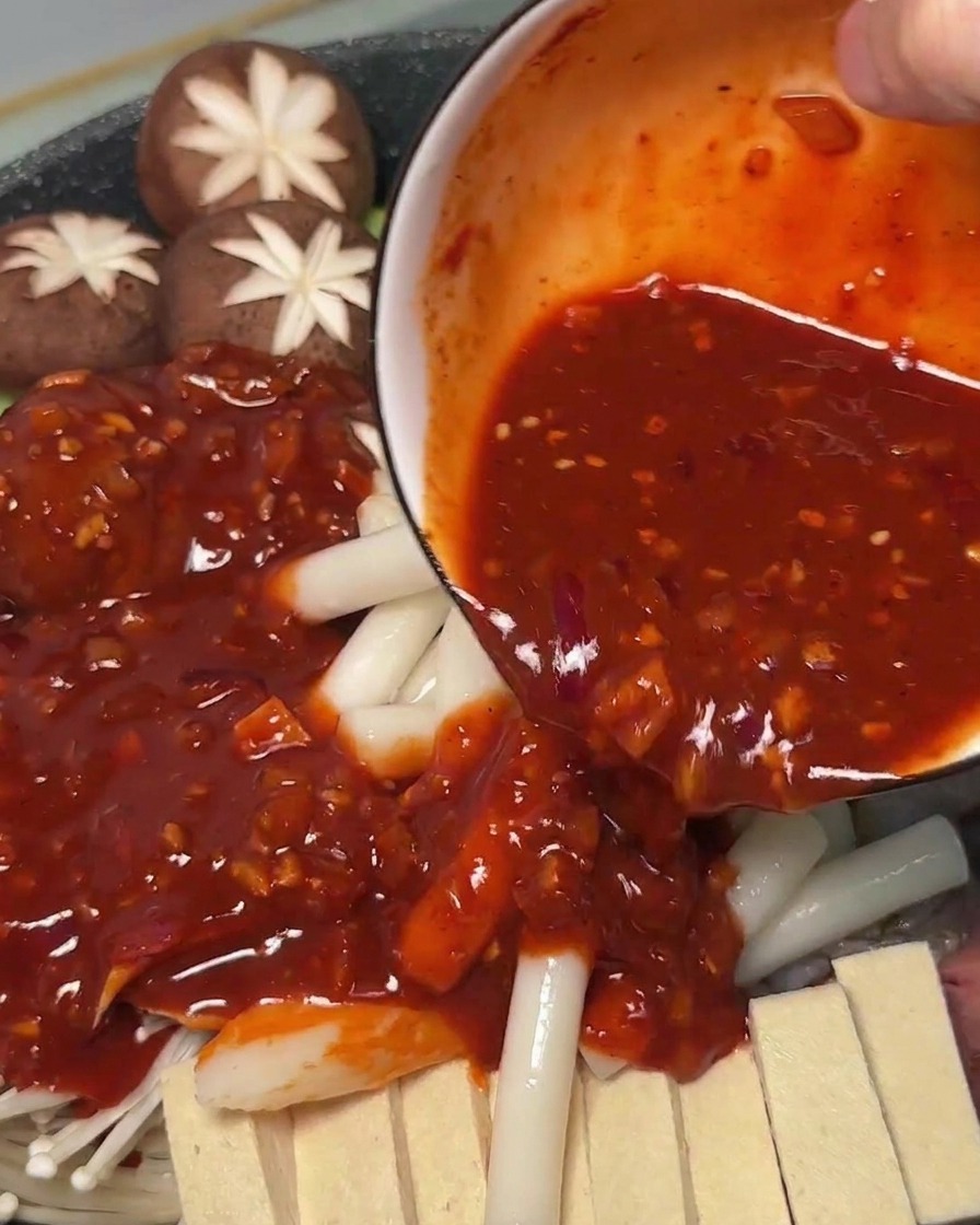 Salsa roja picante vertiéndose sobre los ingredientes crudos y los pasteles de arroz dentro de la olla.