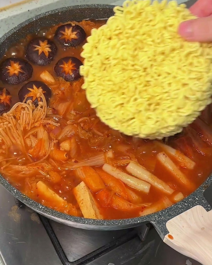 Una mano poniendo un bloque de fideos ramen secos en una olla de sopa roja hirviendo.