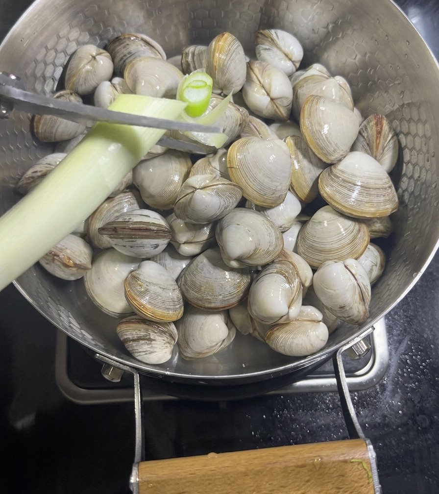 Una mano usando tijeras para cortar una rama de cebolleta sobre una sartén llena de almejas blancas crudas y rodajas de jengibre.