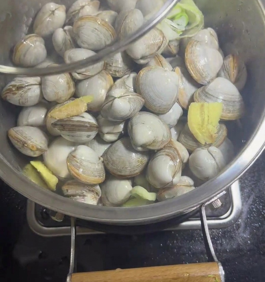 Una sartén de acero inoxidable con almejas y aromáticos siendo cubierta con una tapa de vidrio transparente.