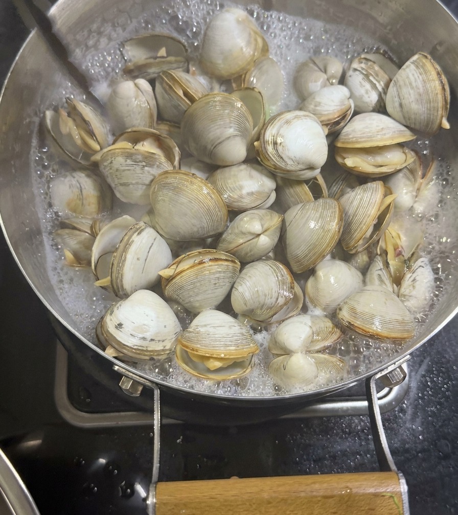 Pinzas metálicas retirando almejas blancas abiertas y cocidas de una sartén con un caldo de mariscos blanquecino y burbujeante.