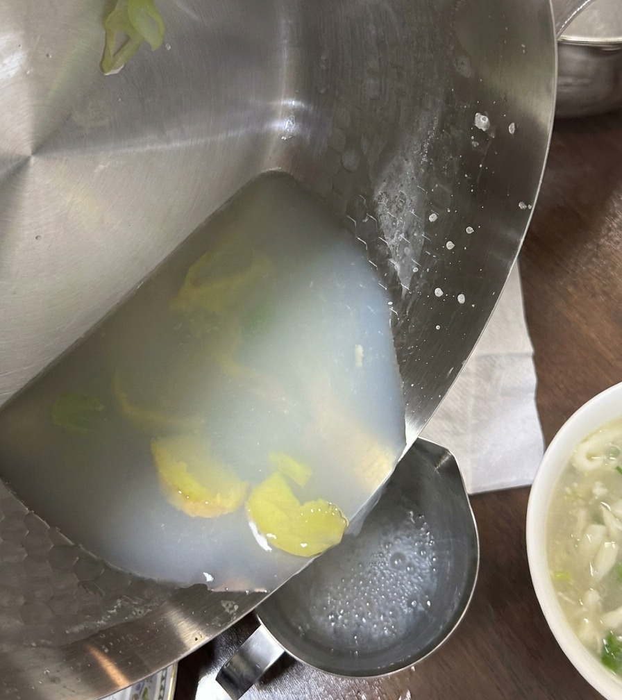 Caldo de almejas blanquecino vertiéndose desde una sartén de acero inoxidable a un recipiente, dejando atrás rodajas de jengibre y sedimentos.