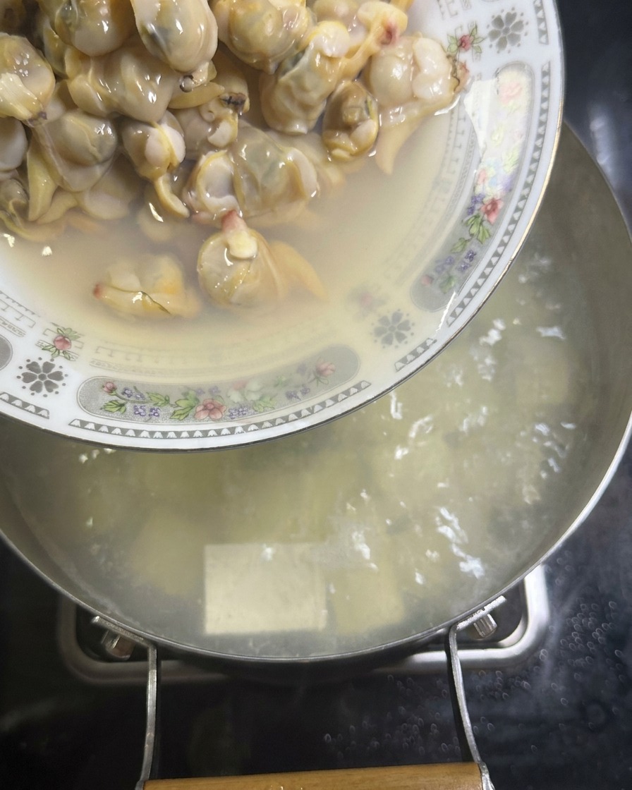 Un plato con carne de almejas ya cocidas se vacía en una olla de caldo blanco hirviendo que contiene cubos de tofu sedoso.