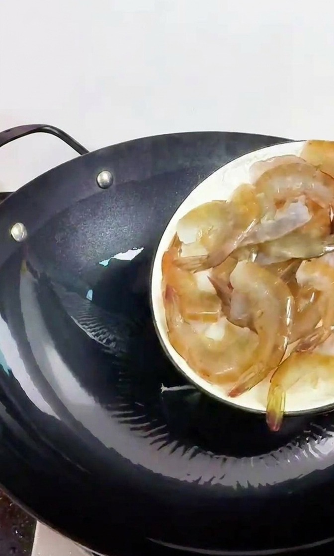 Una mano deslizando gambas frescas y translúcidas en un wok de hierro negro precalentado.
