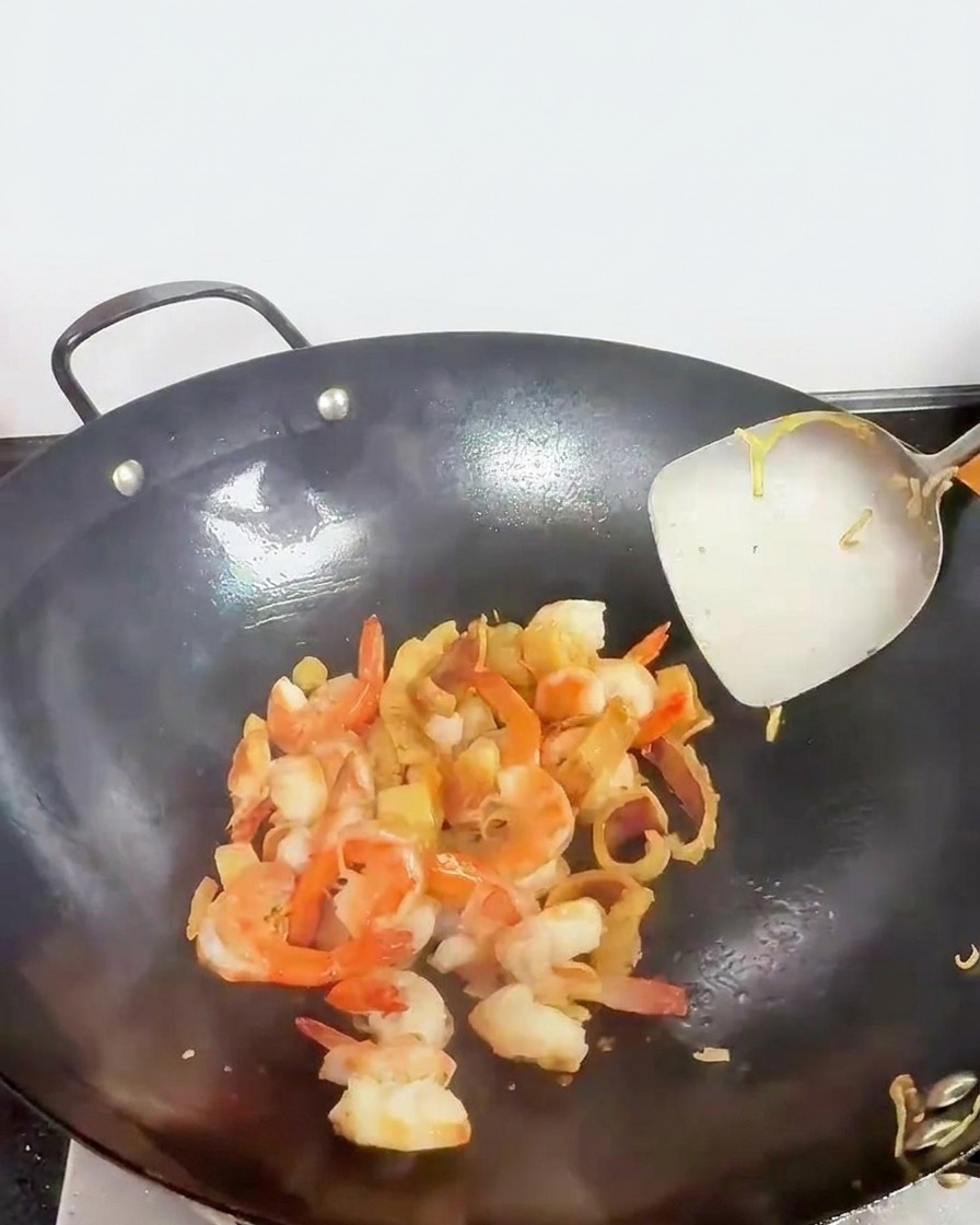 Trozos de marisco, incluyendo gambas rosadas, siendo removidos en un wok con una espátula metálica.