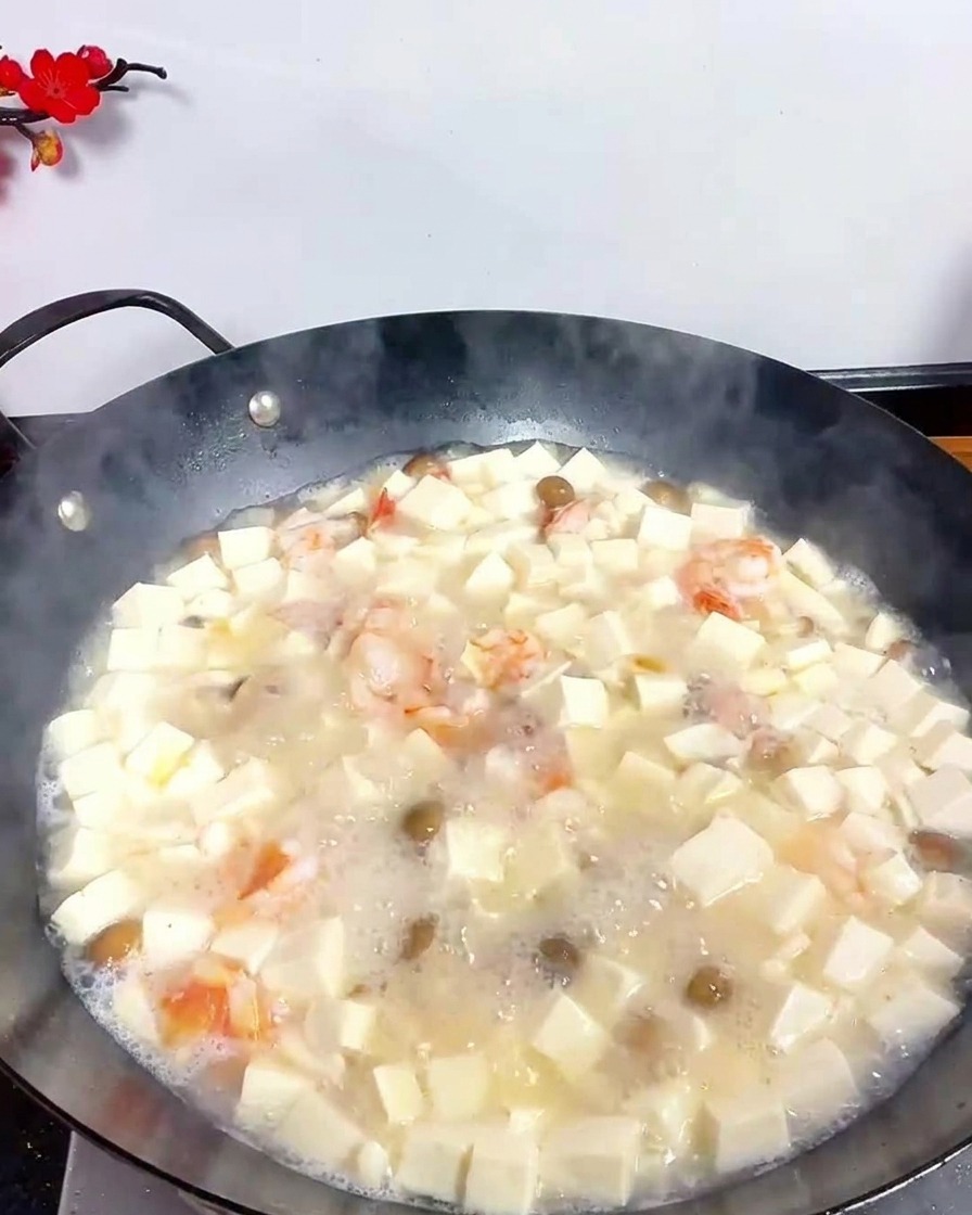 Una cuchara metálica espolvoreando pimienta blanca en una sopa de tofu y mariscos que burbujea en un wok negro.