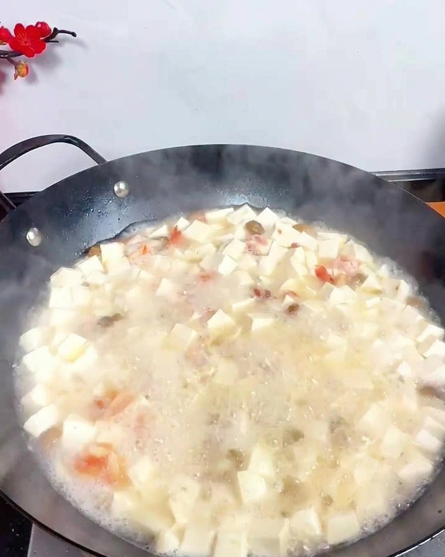 Un cazo de acero inoxidable vertiendo vinagre oscuro en una sopa blanca hirviendo con tofu y mariscos.
