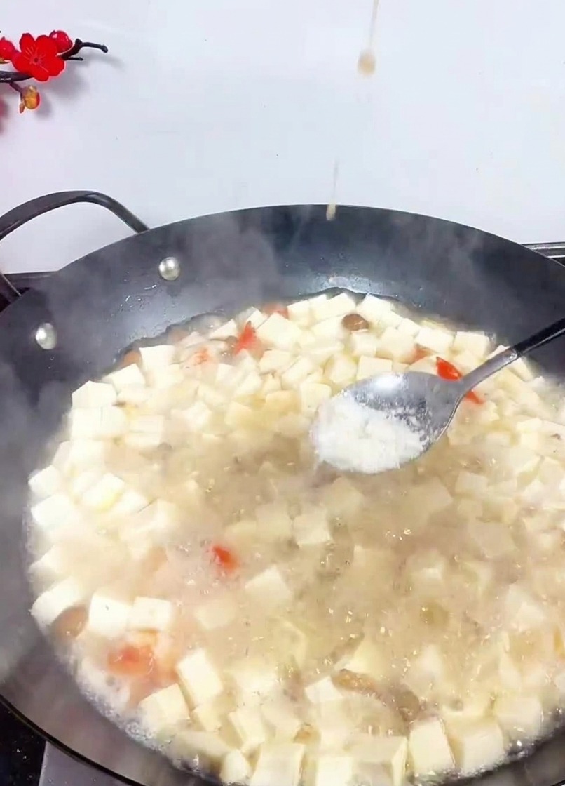 Una cuchara metálica espolvoreando sal blanca en un wok lleno de dados de tofu y mariscos en un caldo ligero.