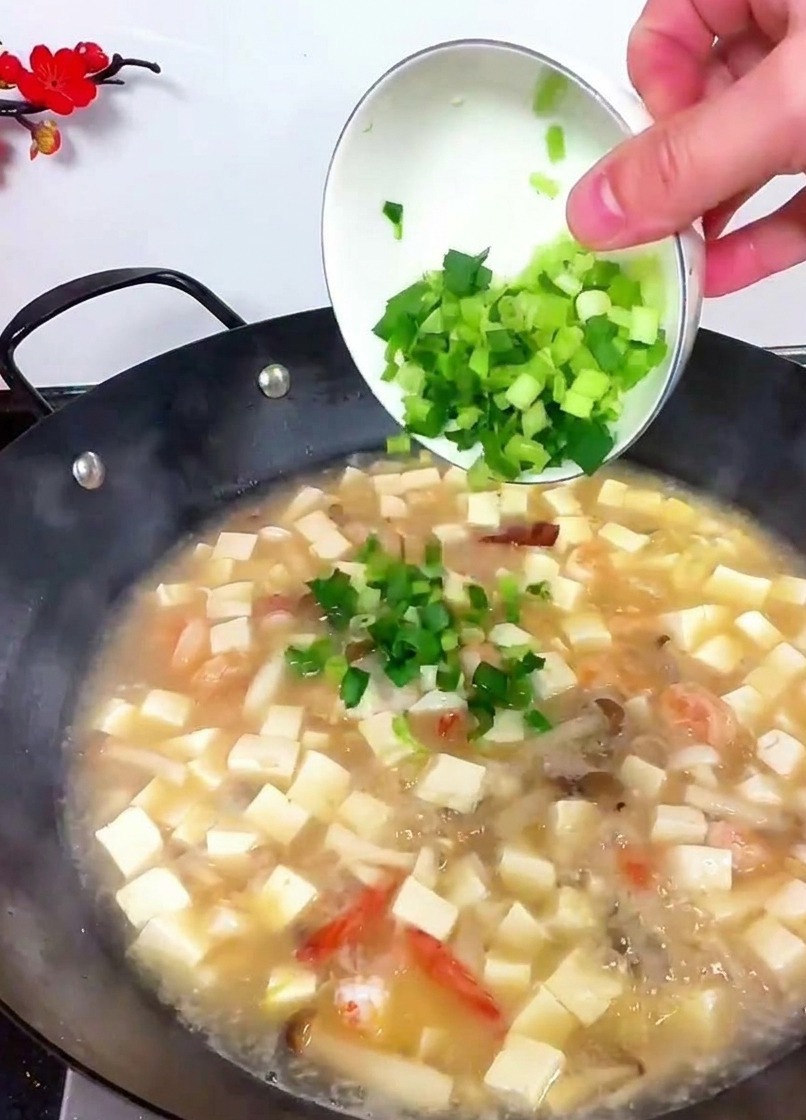 Una mano inclinando un bol blanco para verter apio verde y brotes de ajo en un wok de sopa de tofu.