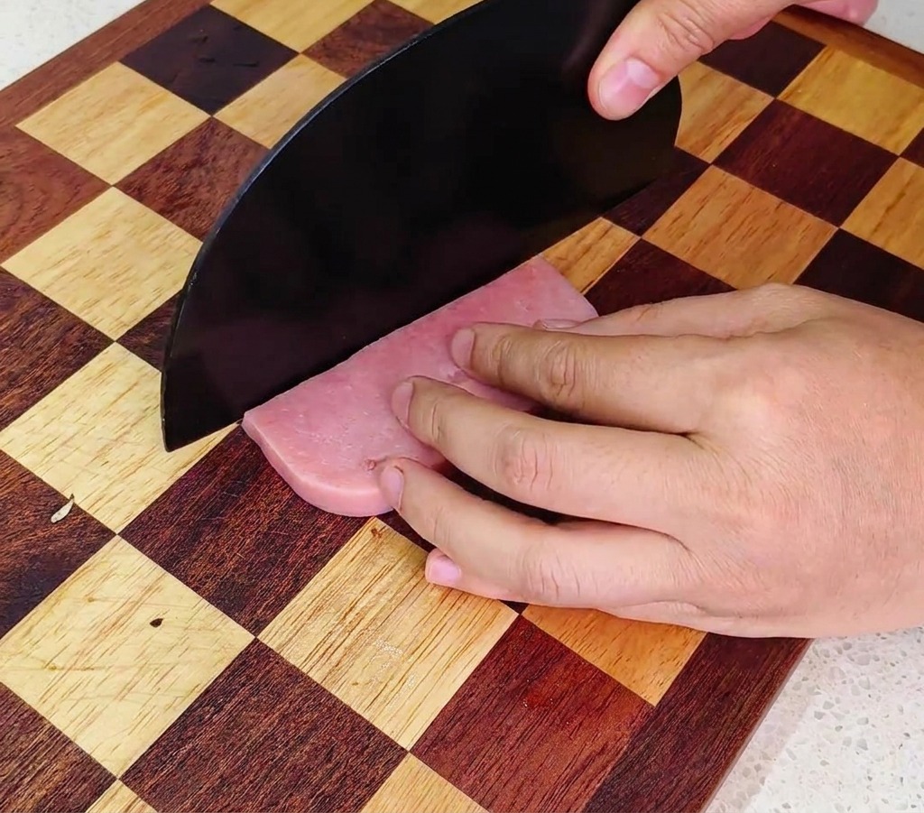 Cuchillo de chef picando una salchicha de jamón rosada en dados pequeños sobre una tabla de madera.