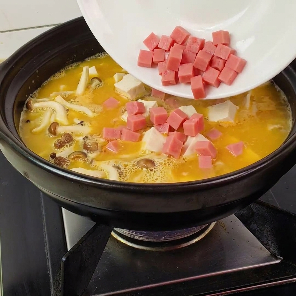 Echando jamón picado, tofu y setas en la olla de barro con el caldo amarillo hirviendo.
