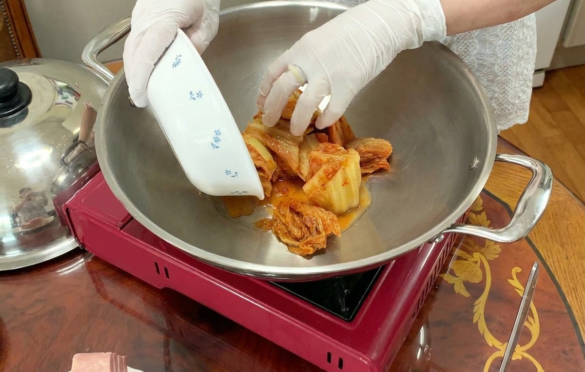 Una persona con guantes blancos coloca kimchi troceado desde un bol pequeño en el centro de una olla grande de acero inoxidable.
