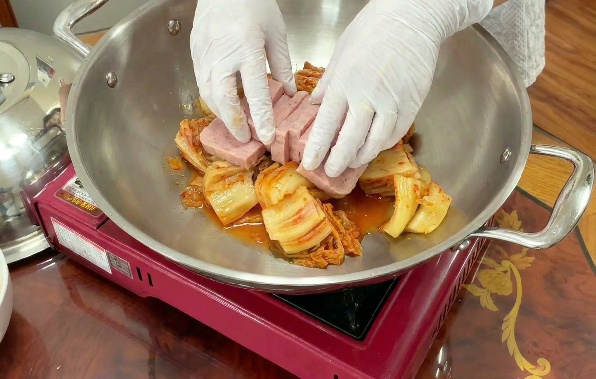 Manos con guantes organizando cuidadosamente rebanadas rectangulares de spam sobre una cama de kimchi en una olla de acero.