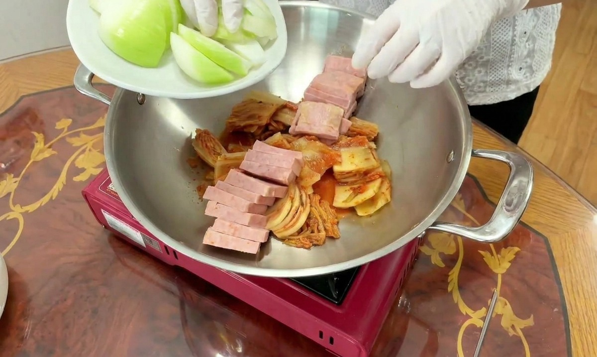 Un cocinero añade gajos de cebolla blanca fresca a una olla que ya contiene kimchi y spam laminado.