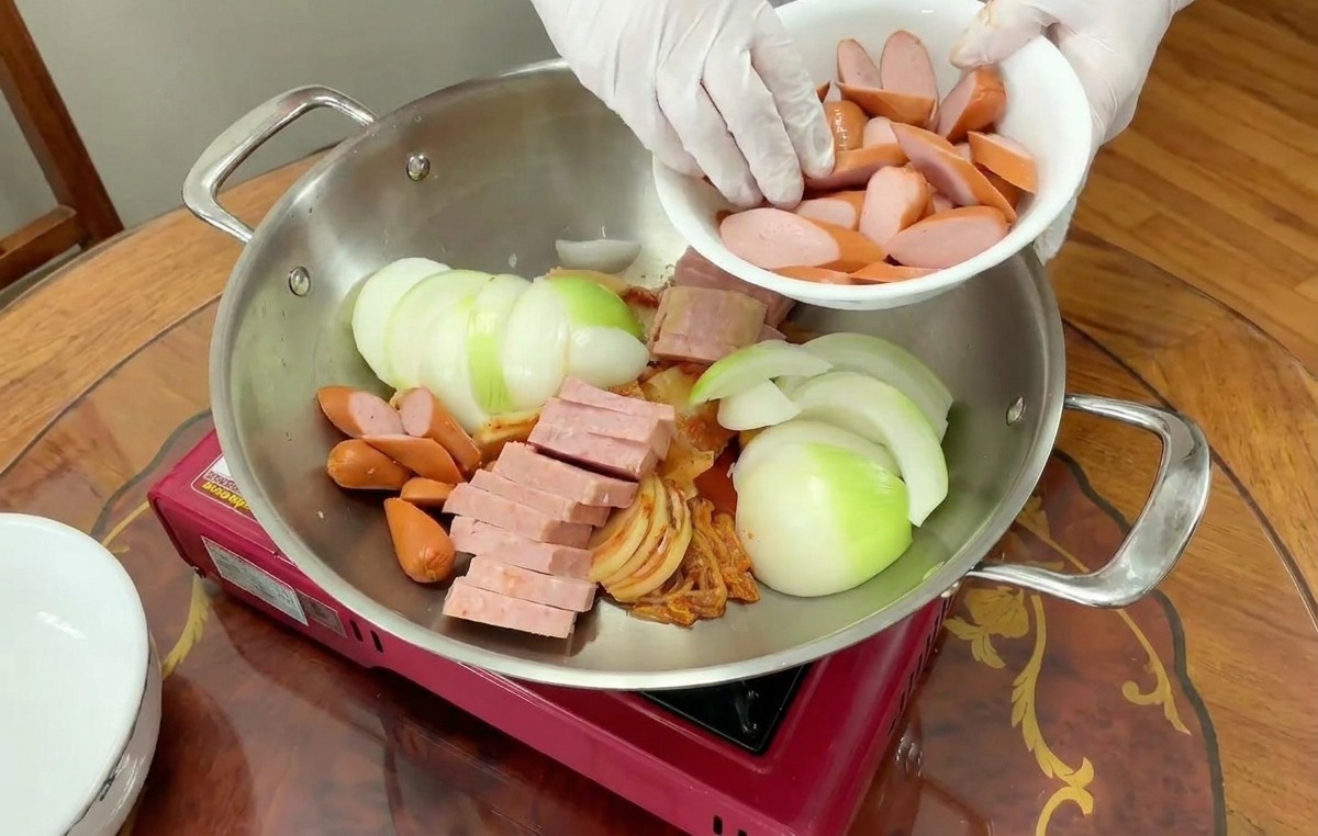 Salchichas troceadas cayendo desde un bol blanco hacia una olla con cebolla, jamón y kimchi.