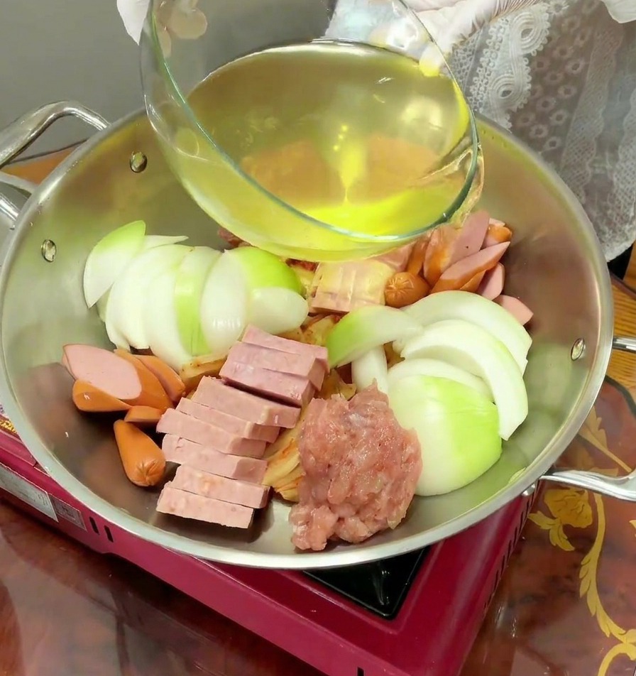 Un líquido amarillento claro se vierte desde un bol de cristal a una olla de acero llena de ingredientes crudos.
