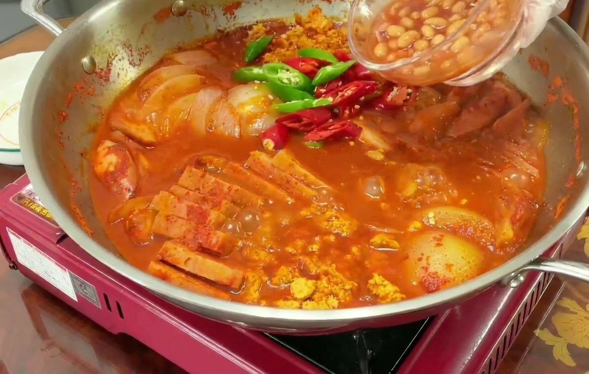 Una mano vertiendo frijoles blancos en salsa desde un bol pequeño a una olla hirviendo que ya tiene chiles verdes y rojos.