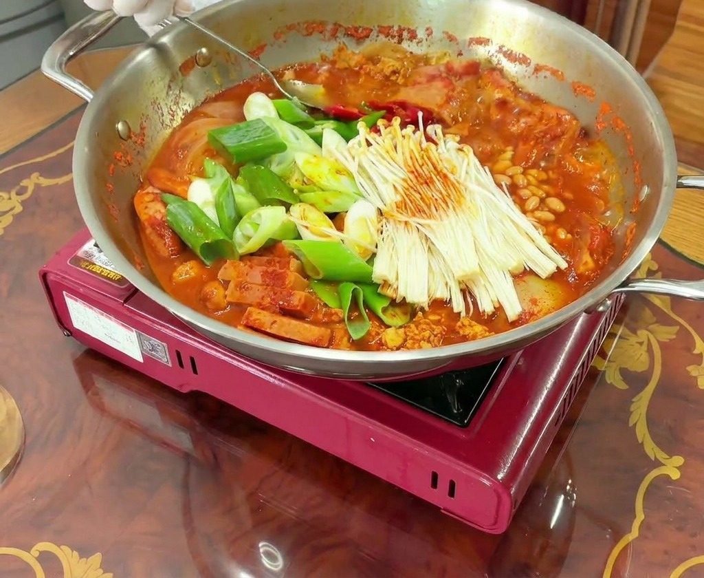 Manos con guantes usando una cuchara para remover una olla burbujeante de Budae Jjigae, mezclando la carne y las verduras con el caldo rojo.