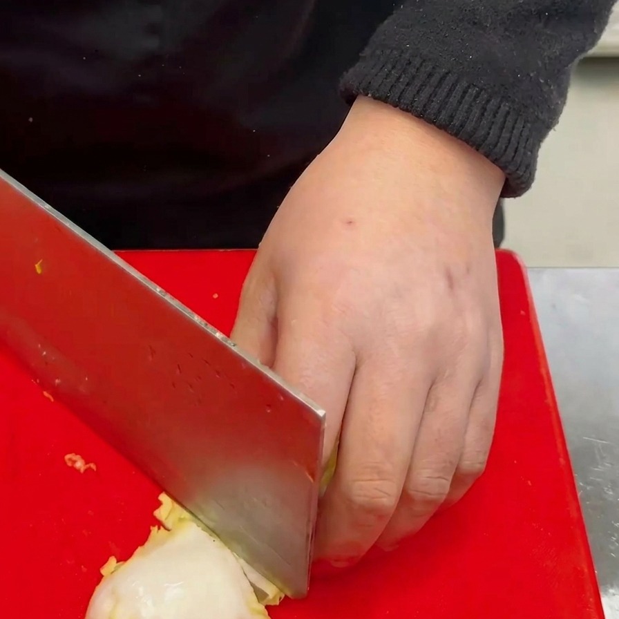 Una mano usando un cuchillo grande para cortar una pequeña col blanca en trozos gruesos sobre una tabla de cortar roja.